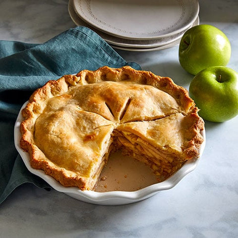 Apple pie in a stoneware pie baking dish with slice removed, along with green apples and teal napkin close.