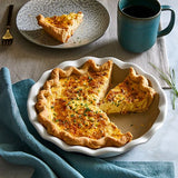 Quiche pie in a stoneware pie baking dish with a slice removed on a plate, next to a blue mug and a teal napkin.